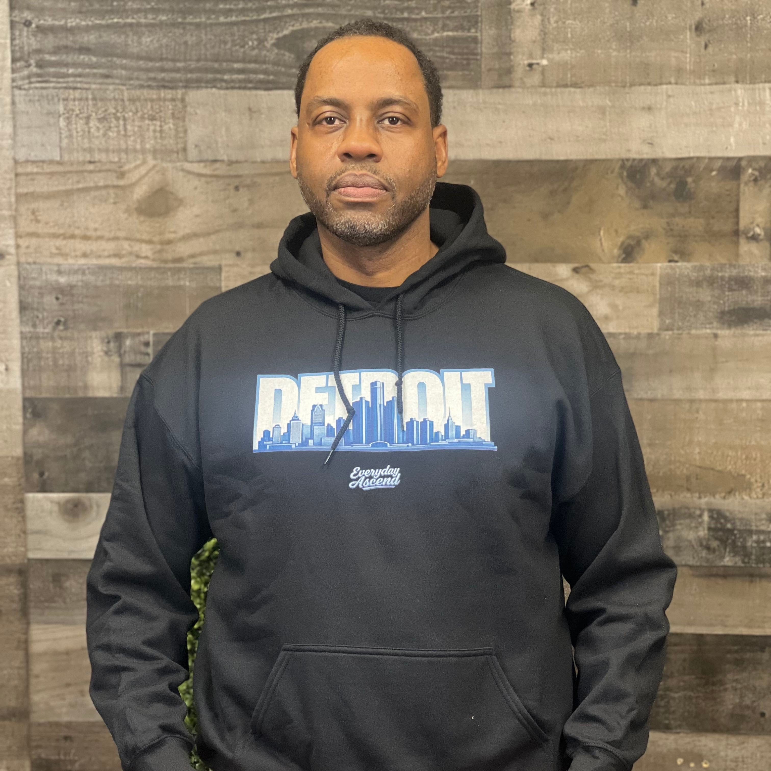 Man wearing a black hoodie with 'Detroit Skyline Everyday Ascend printed on it against a wooden paneled wall.