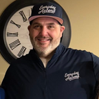 Man wearing a navy blue quarter zip and cap with 'Everyday Ascend' branding, standing in front of a decorative clock.